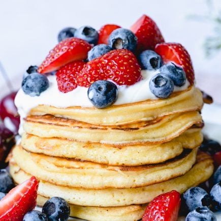Une pile de pancakes sans levure chimique saines garnies de crème fouettée, de bleuets frais et de fraises tranchées sur une assiette blanche.