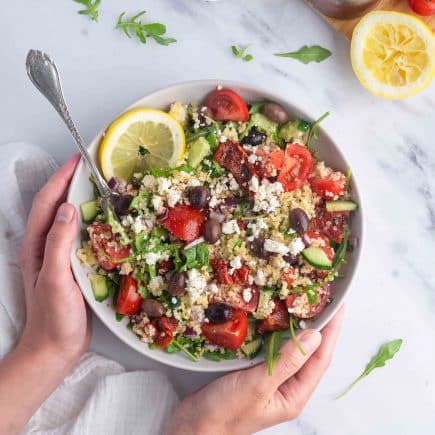 Hands holding a bowl of bulgur salad with ingredients including quinoa, cherry tomatoes, cucumbers, olives, feta cheese, and a lemon wedge; herbs and dressing in the background on a white marble surface.