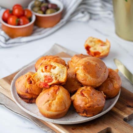 Une assiette de Muffins Salés Végétariens avec tomates hachées visibles et fromage, posée sur une planche de bois. En arrière-plan, on aperçoit des bols de tomates cerises, des olives et une plante verte.