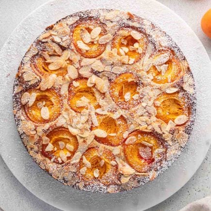 Un gâteau rond abricot et amandes sans gluten, garni de tranches d'amandes et de sucre en poudre, est présenté sur une surface blanche, avec des abricots frais et une cuillère à proximité.