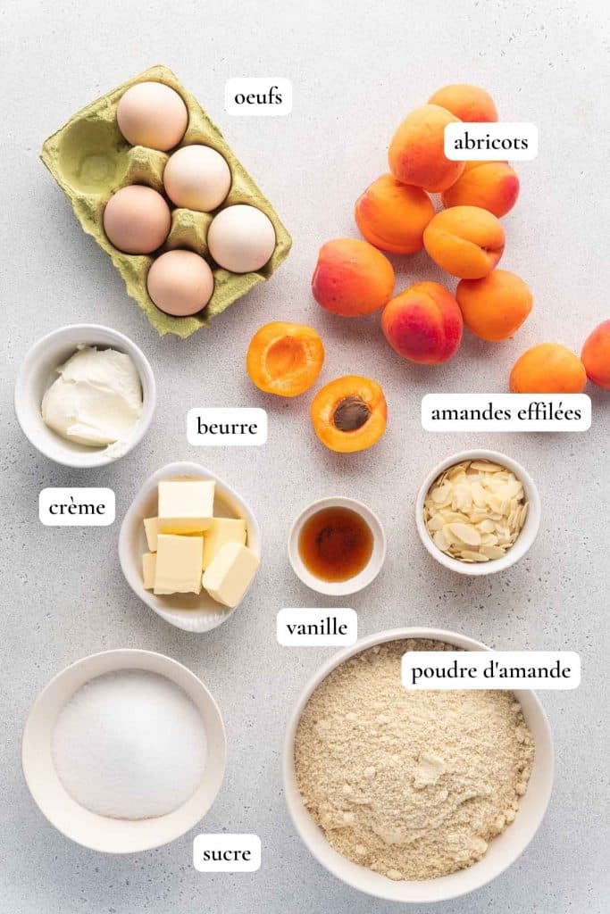 Les ingrédients d'une recette de gâteau aux abricots et amandes sont affichés sur une surface, notamment les œufs, les abricots, le beurre, la crème, les amandes effilées, la vanille, la poudre d'amandes et le sucre