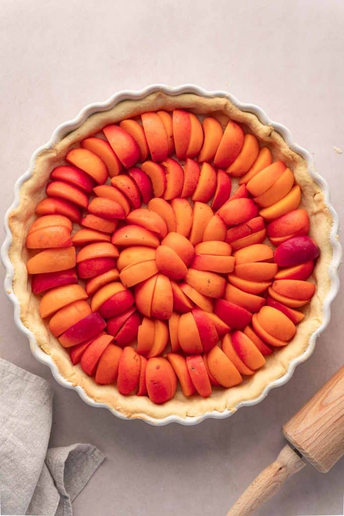 Vue de dessus d'une tarte non cuite avec des tranches d'abricots soigneusement disposées selon un motif circulaire, placées sur une surface de couleur claire à côté d'un rouleau à pâtisserie et d'un tissu gris.
