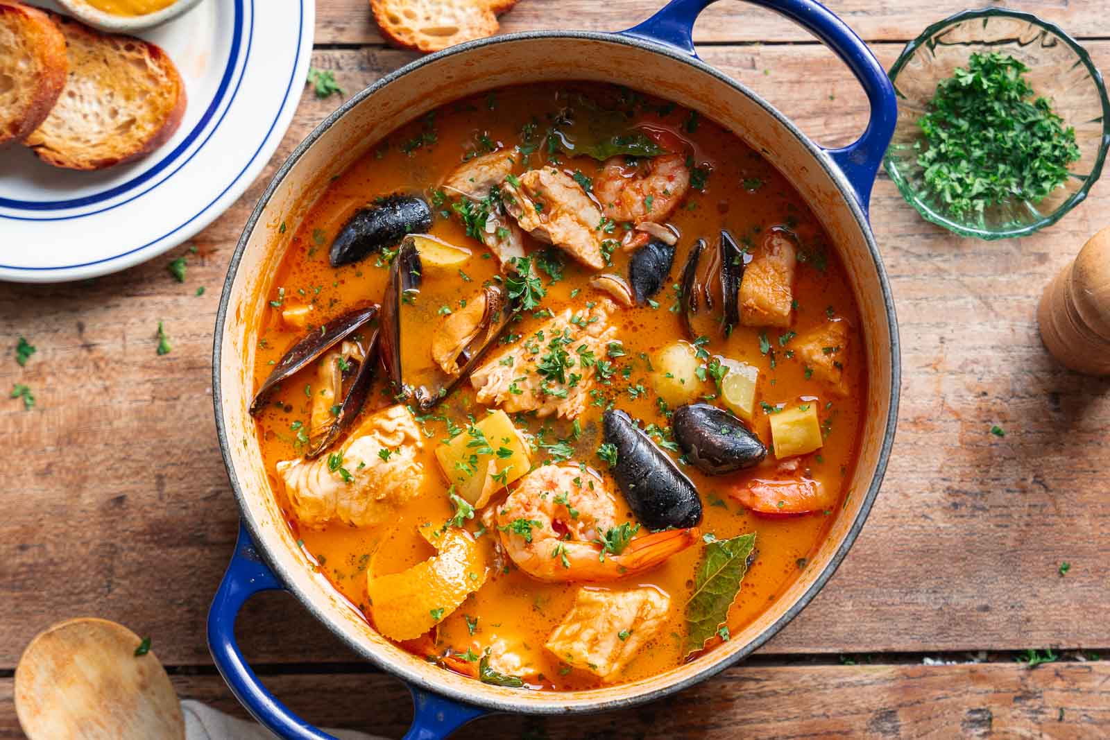 Une casserole de bouillabaisse, une soupe de poisson française traditionnelle regorgeant de crevettes, de poisson, de moules, de pommes de terre et d'herbes aromatiques, trône sur une table en bois à côté d'un bol de persil haché et de tranches de pain grillé.