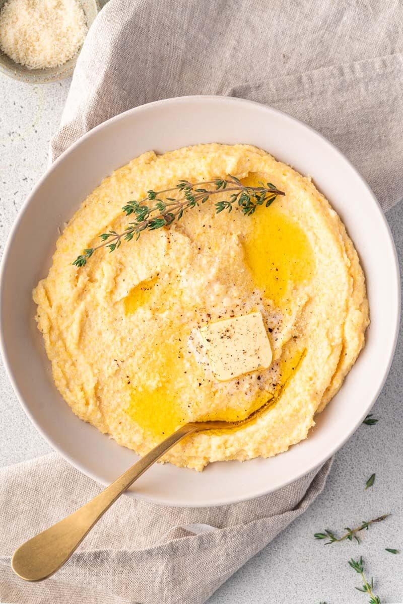 A bowl of creamy parmesan polenta topped with melting butter, fresh thyme, and black pepper, with a spoon resting inside the bowl.