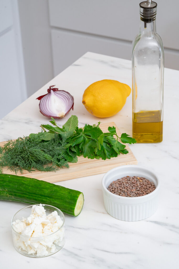 Zutaten auf einer Küchentheke, um einen zuzubereiten: eine halbe rote Zwiebel, eine Zitrone, eine Flasche Olivenöl, frische Kräuter auf einem Schneidebrett, Gurke für den Linsensalat mit Minze und Feta, Fetakäse in einer Schüssel und goldene Linsen in einem Auflaufförmchen.