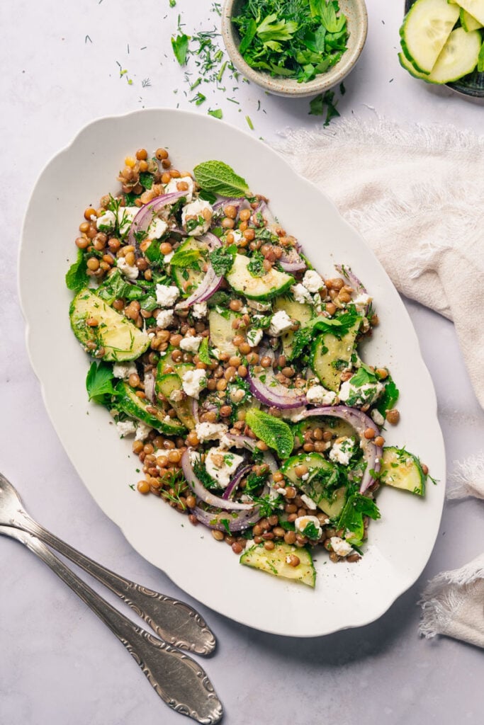 Une assiette ovale de salade lentilles, menthe et feta avec des tranches de concombres, des oignons rouges, du fromage feta et des herbes fraîches. Un petit bol d'herbes hachées et une serviette en tissu blanc se trouvent à côté de l'assiette.