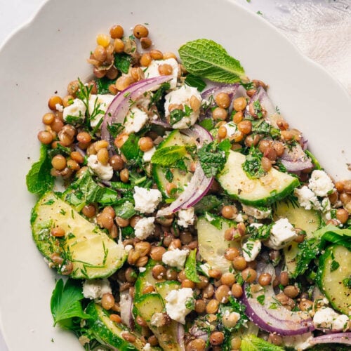 Un plat blanc avec Salade lentilles menthe feta garnie de tranches de concombre, d'oignon rouge, de fromage feta, d'herbes fraîches. Un petit bol d’herbes hachées se trouve en arrière-plan.