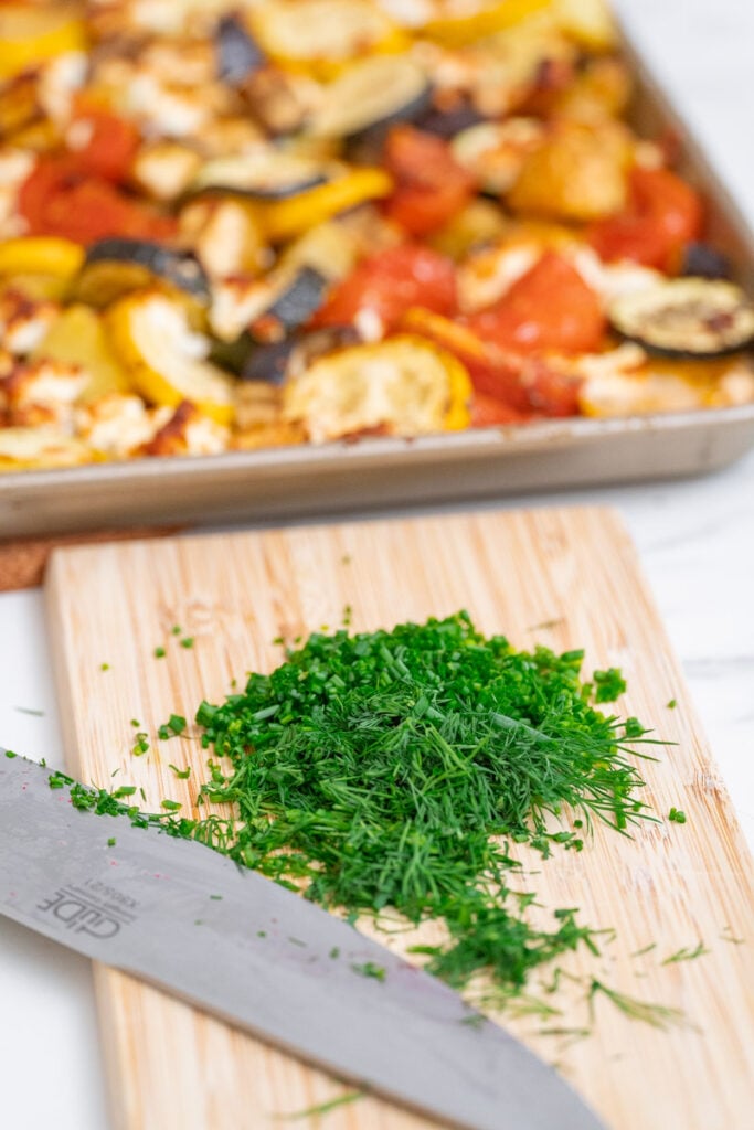 Chopped fresh herbs on a wooden cutting board next to a kitchen knife, with a tray of roasted vegetables in the background.