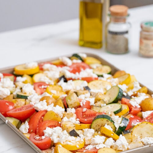 A baking sheet lined with a mixture of chopped zucchini, tomatoes, yellow squash and crumbled cheese. Two spice jars and a bottle of olive oil are visible in the background.