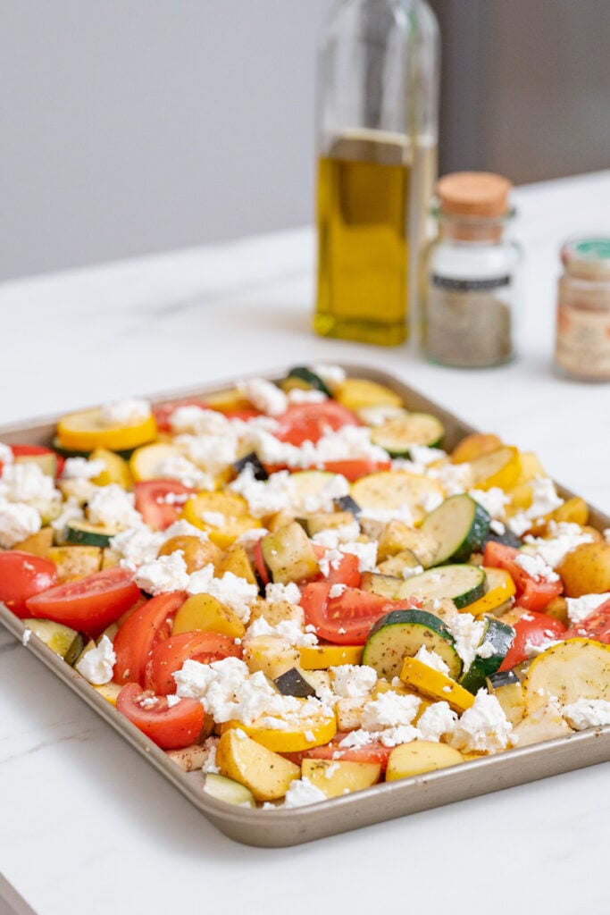 A baking sheet lined with a mixture of chopped zucchini, tomatoes, yellow squash and crumbled cheese. Two spice jars and a bottle of olive oil are visible in the background.