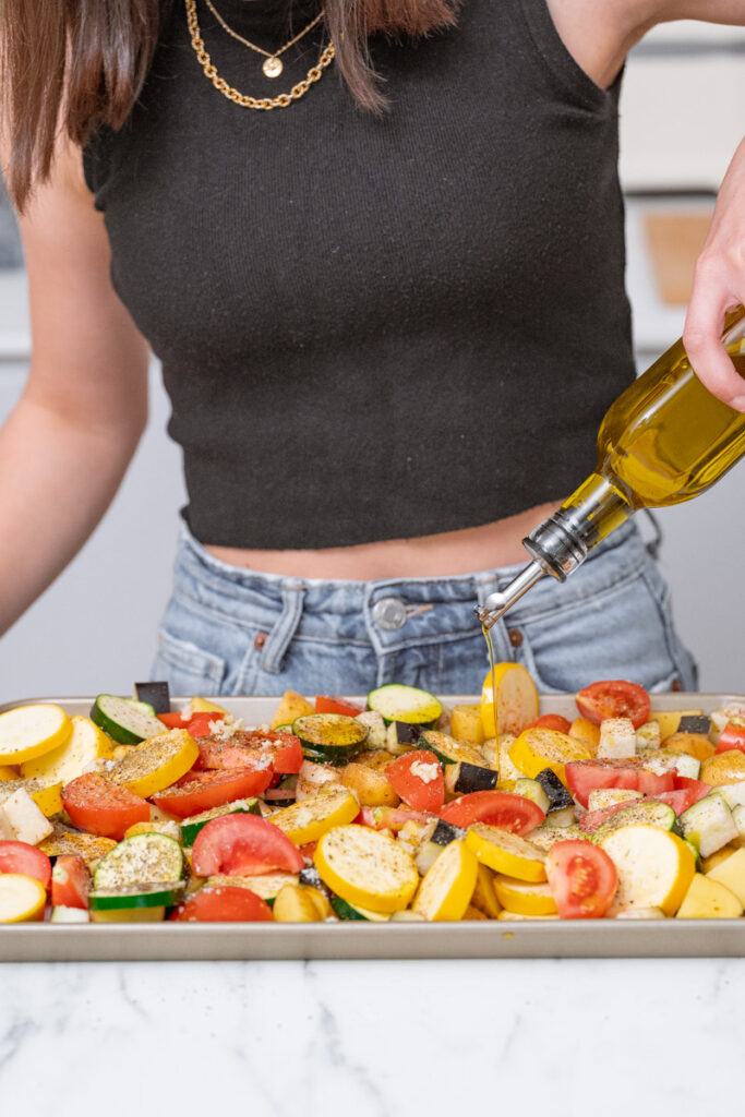 A person dressed in a black top and jeans pours olive oil onto a tray of sliced vegetables, including tomatoes, zucchini and yellow squash, in a kitchen.