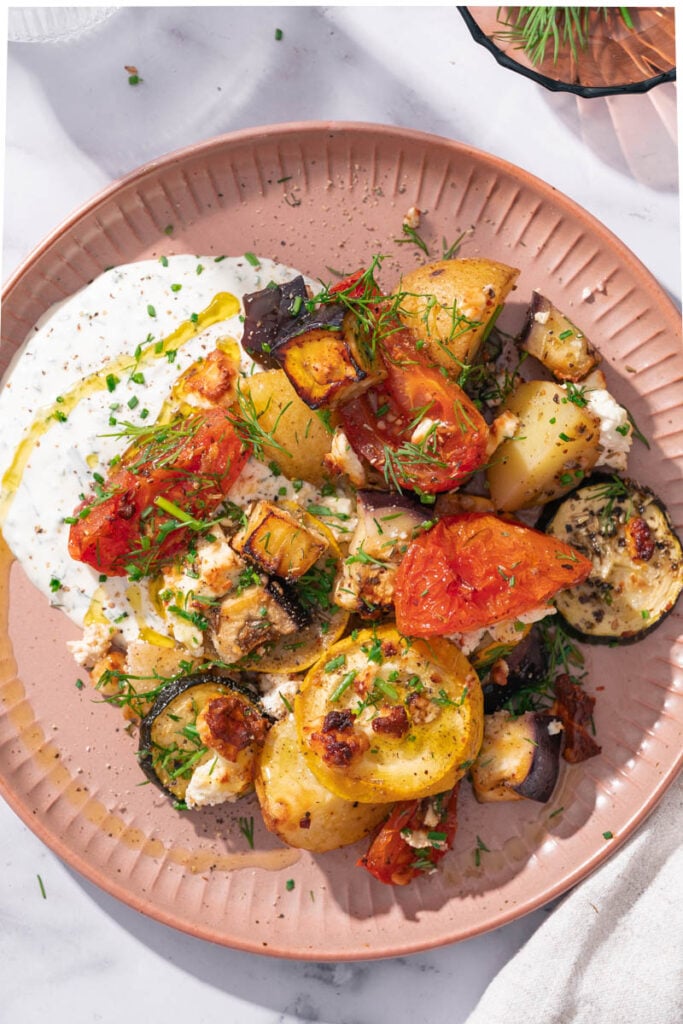 A plate of roasted vegetables including tomatoes, zucchini and potatoes on a bed of creamy sauce, garnished with herbs.