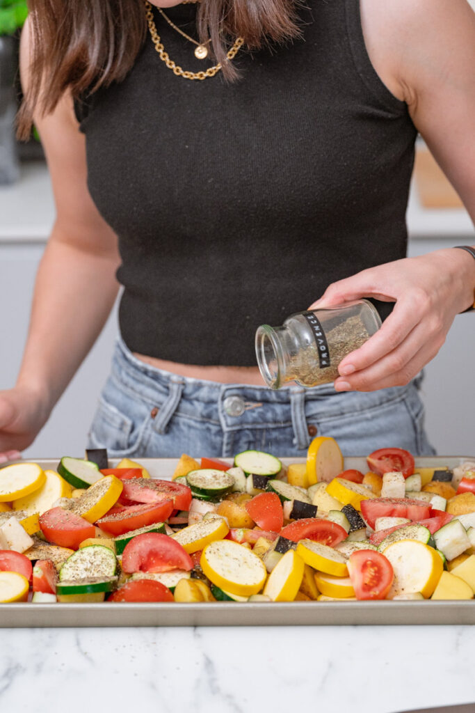 A person dressed in a sleeveless black top is seasoning a tray of sliced vegetables with herbs from a jar in a kitchen.