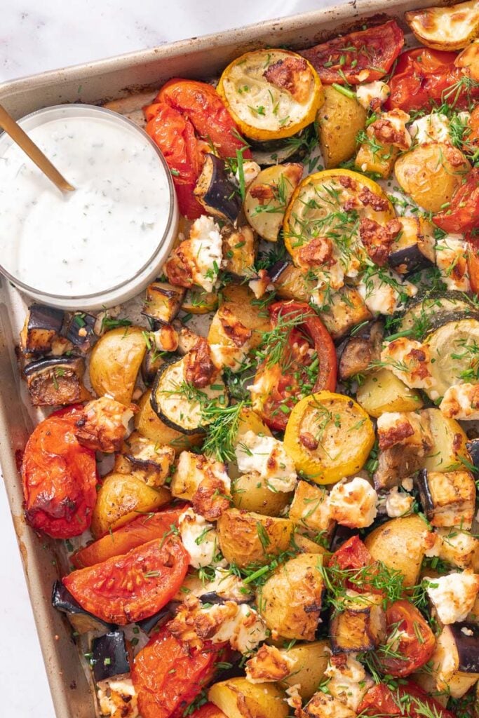 A baking sheet filled with roasted vegetables, including tomatoes, zucchini and potatoes, and topped with herbs and feta cheese. A small bowl of creamy dip with a spoon is placed on the tray.