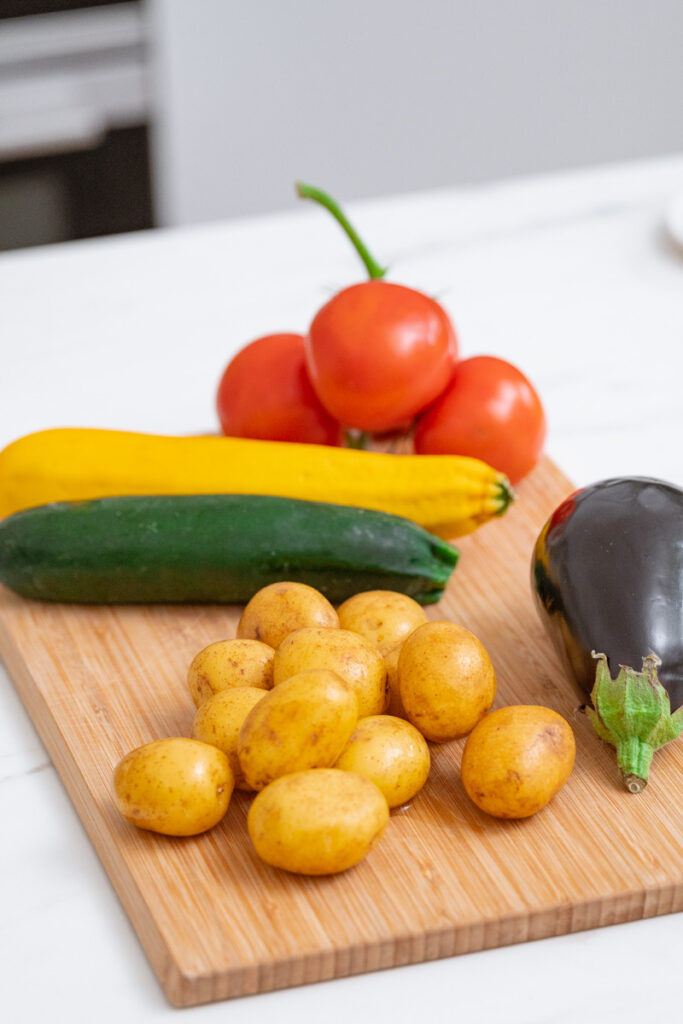A wooden cutting board with vegetables: small yellow potatoes, an eggplant, a green zucchini, a yellow zucchini and three red tomatoes.