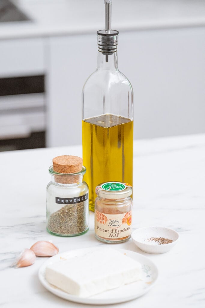 A bottle of olive oil, a jar of herbes de Provence, a jar of piment d'Espelette, two cloves of garlic, a block of feta cheese on a plate and a small dish of ground pepper on a marble counter.