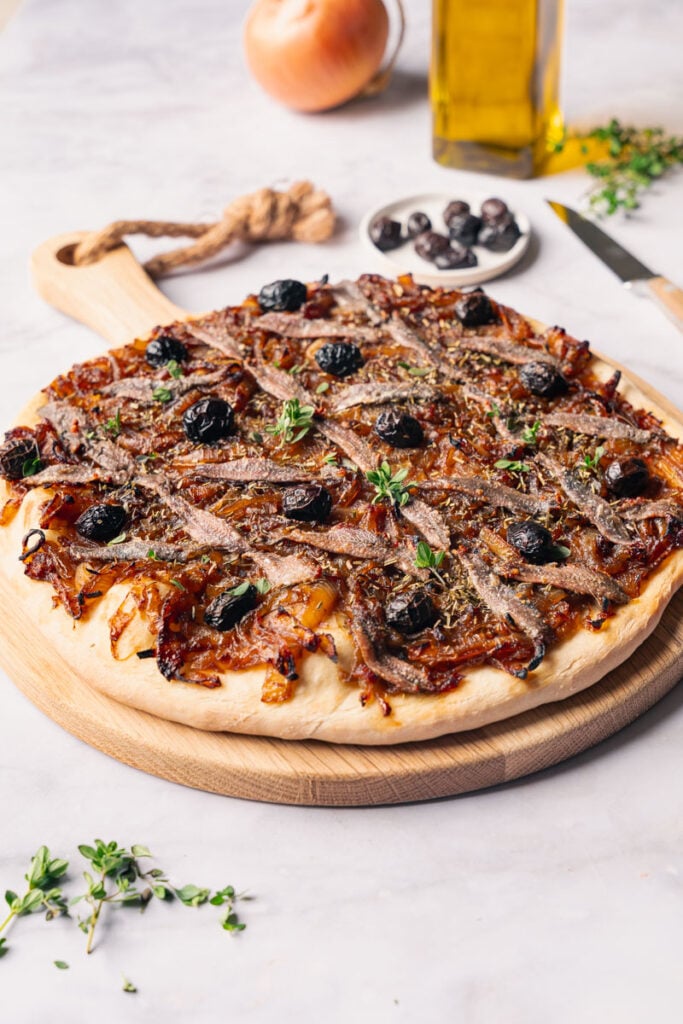 Une pissaladière niçoise fraîchement cuite, garnie d'anchois, d'olives noires, d'herbes et d'oignons caramélisés sur une planche en bois.