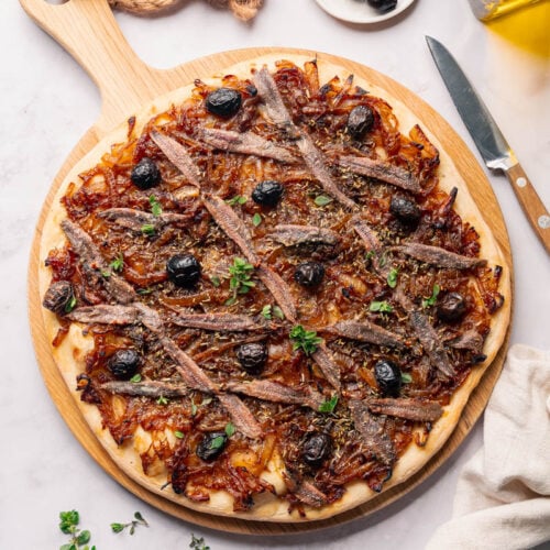 Une pissaladière niçoise préparée garnie d'anchois, d'olives noires et d'oignons caramélisés, servie sur une planche à pizza en bois.