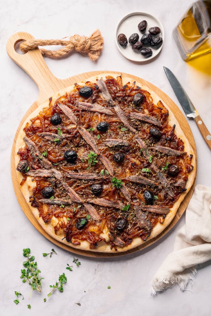 Une pissaladière niçoise préparée garnie d'anchois, d'olives noires et d'oignons caramélisés, servie sur une planche à pizza en bois.