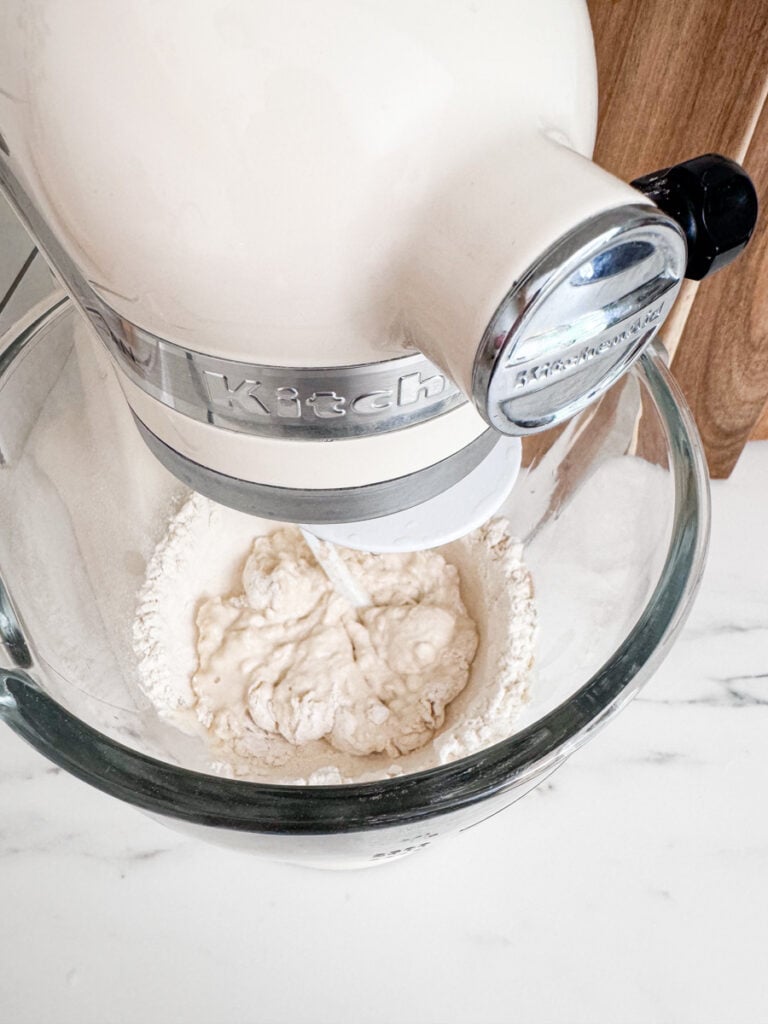 An electric stand mixer with a metal bowl mixes the dough ingredients on a white marble countertop.