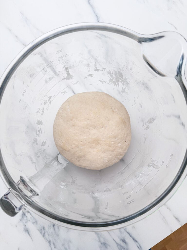 A ball of pizza dough rests in a clear glass bowl on a marble countertop.