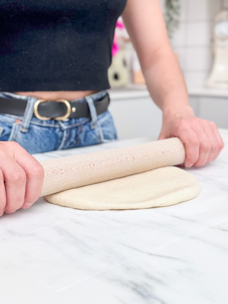 A person wearing a black top and jeans uses a rolling pin to flatten pizza dough to make a niçoise pissaladière.
