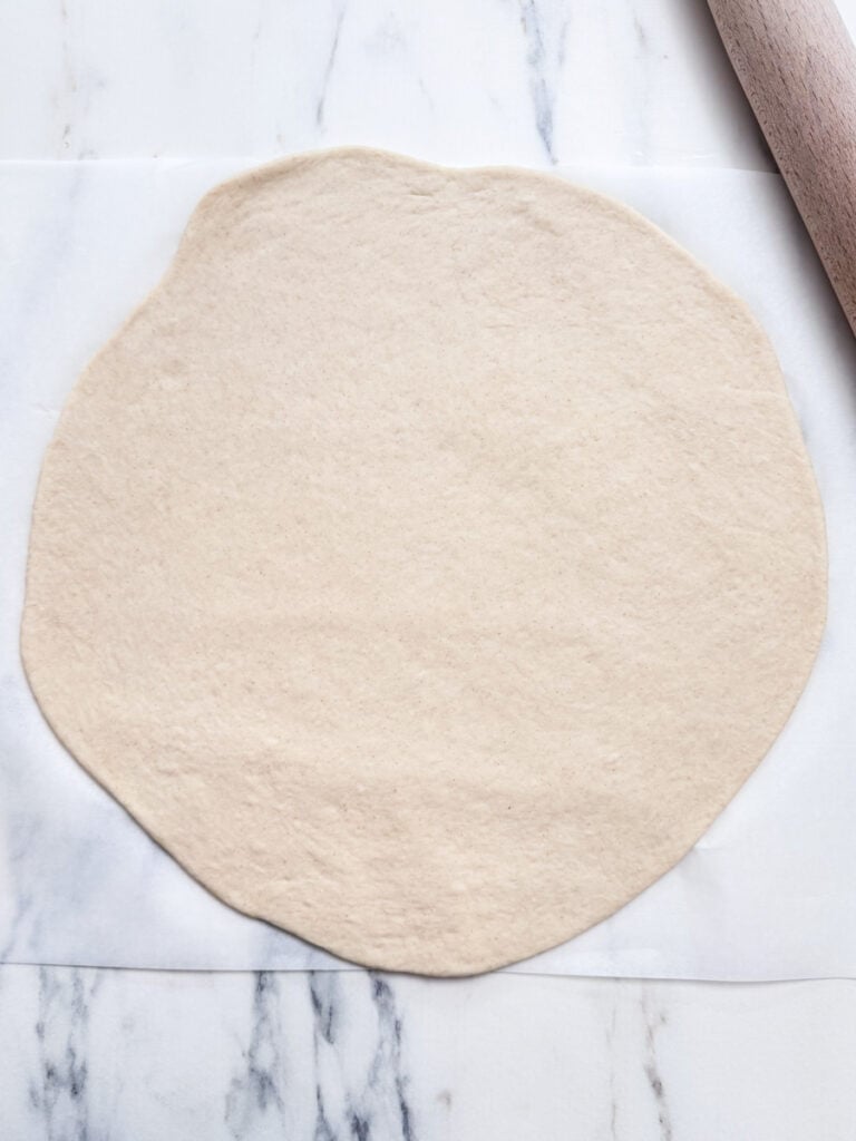 A round, rolled-out dough on a marble surface, with a wooden rolling pin partially visible.