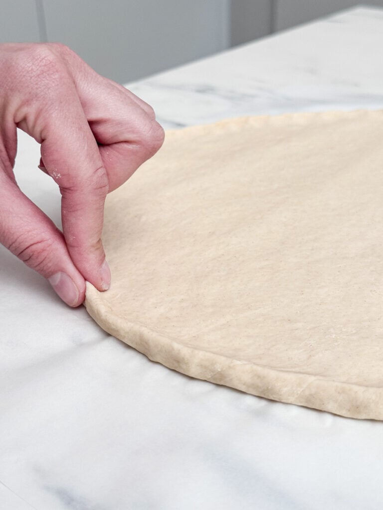 One hand pinches the edge of a pizza dough spread on a marble surface.