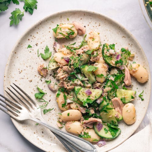 A salad plate of white beans, cucumbers, tuna, red onion and parsley, with two forks on one side.