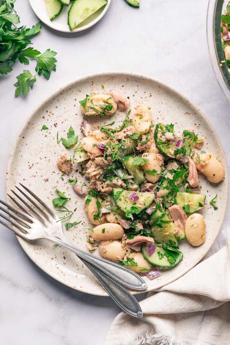 A salad plate of white beans, cucumbers, tuna, red onion and parsley, with two forks on one side.