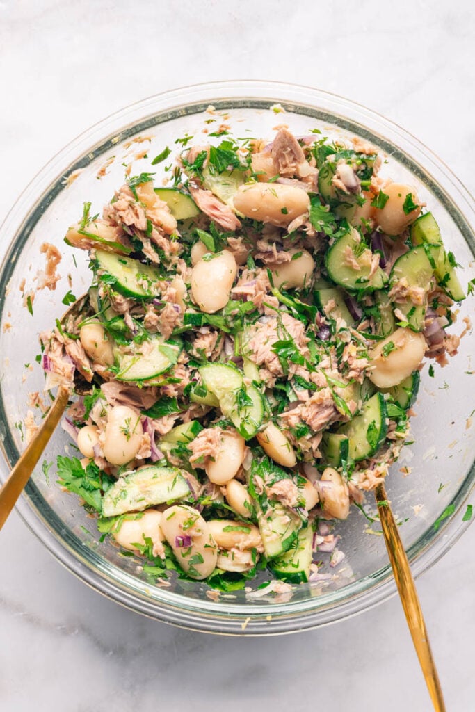 A glass bowl filled with a mixture of white beans, cucumber slices, tuna, red onions and fresh herbs, tossed in a light vinaigrette.