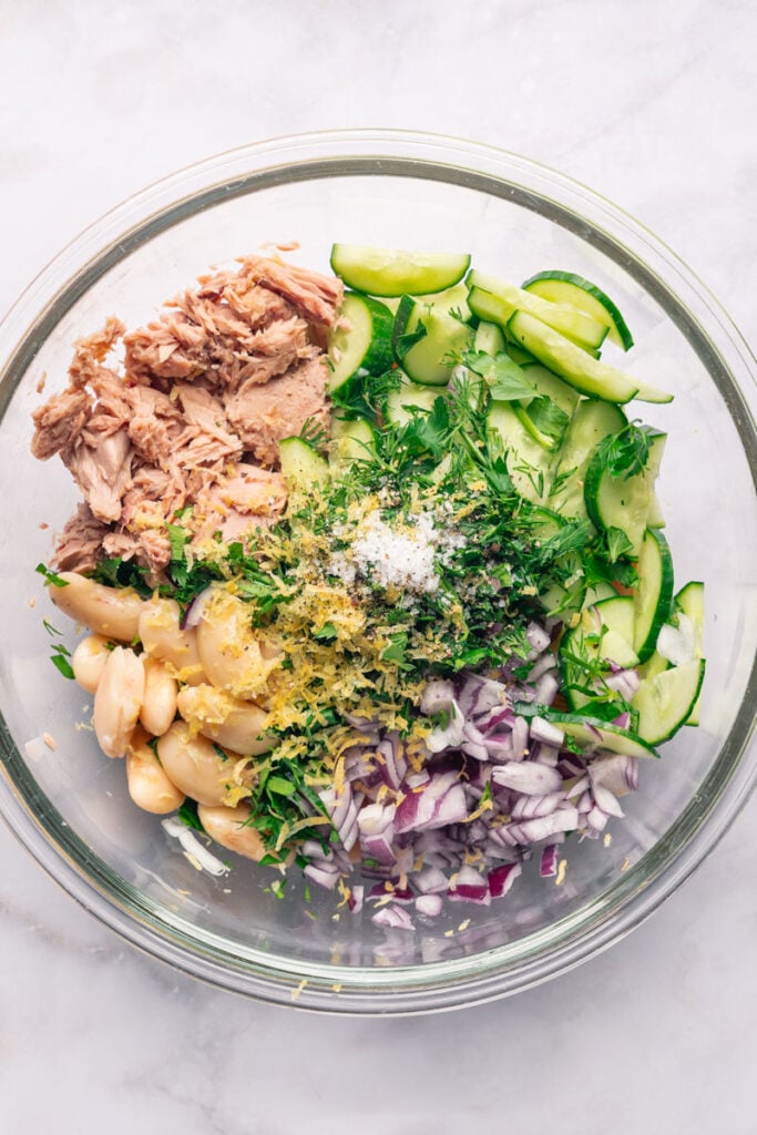 A glass bowl containing salad ingredients, including tuna, cucumber slices, white beans, red onion, chopped herbs, lemon zest and salt.