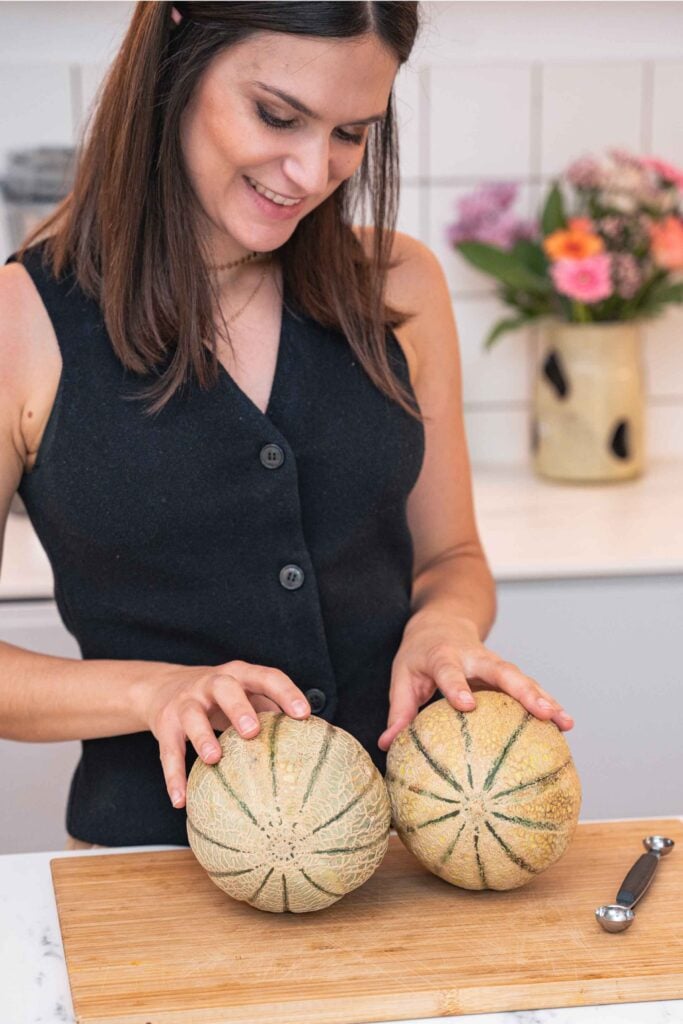 Eine Person in einem schwarzen, ärmellosen Oberteil steht vor einer Küchentheke und hält zwei Cantaloupe-Melonen auf einem Schneidebrett, mit Blumen in einer Vase im Hintergrund.