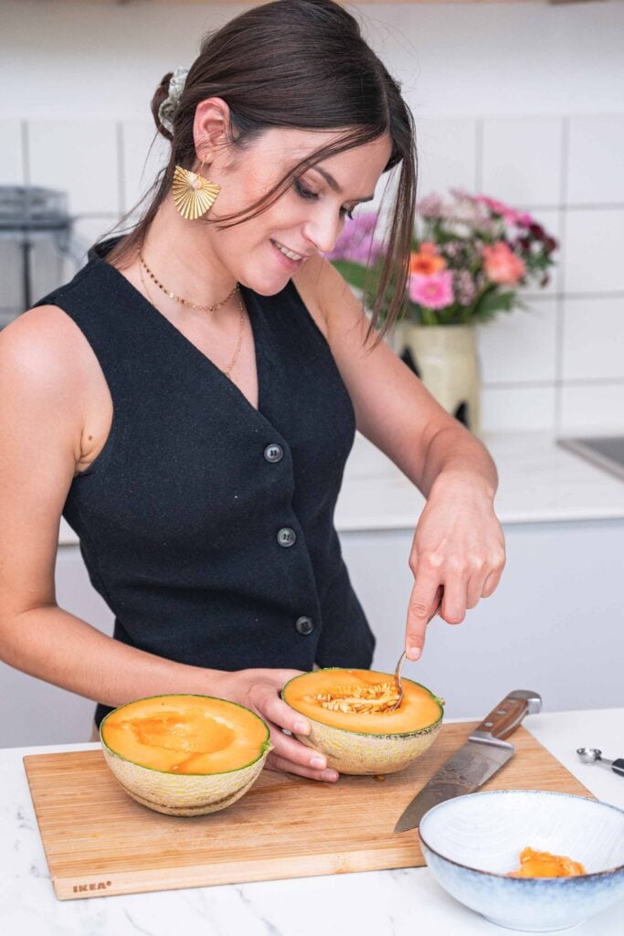 A woman wearing a black sleeveless top scoops seeds from a halved cantaloupe with a spoon in a kitchen. A vase of flowers is in the background. 