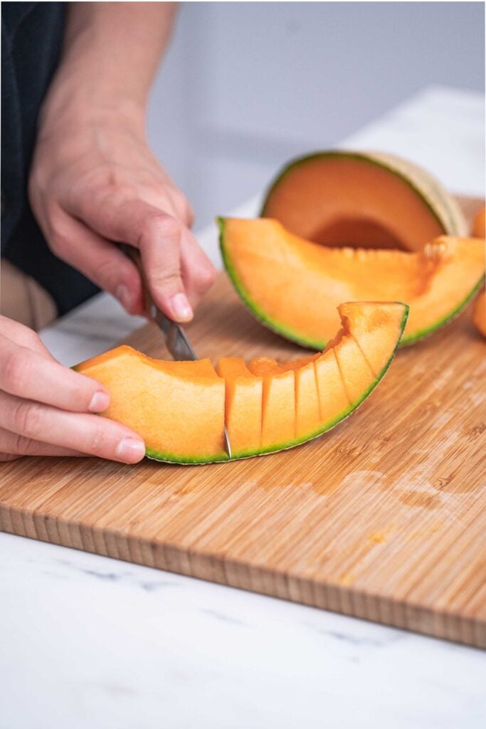 Close-up of a person cutting a cantaloupe on a wooden cutting board with a knife. Several slices and a cantaloupe cut in half are visible in the background. 