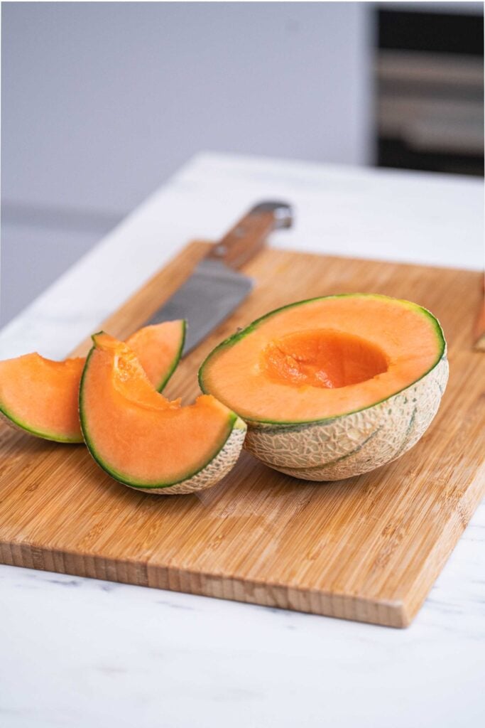 Un cantaloup coupé en deux avec les graines enlevées et deux tranches à côté sur une planche à découper en bois, à côté d'un couteau.