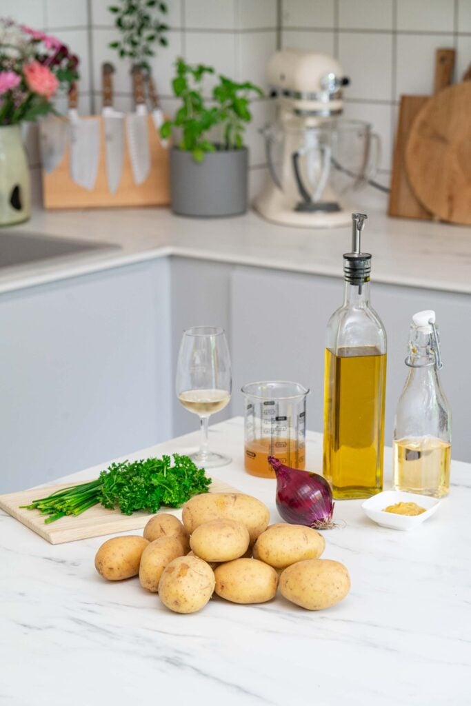 Eine Küchentheke mit Kartoffeln, einer roten Zwiebel, Petersilie auf einem Schneidebrett, einem Messbecher, einem Glas Weißwein, Olivenöl, einer kleinen Schüssel mit Senf und einer Flasche Essig.