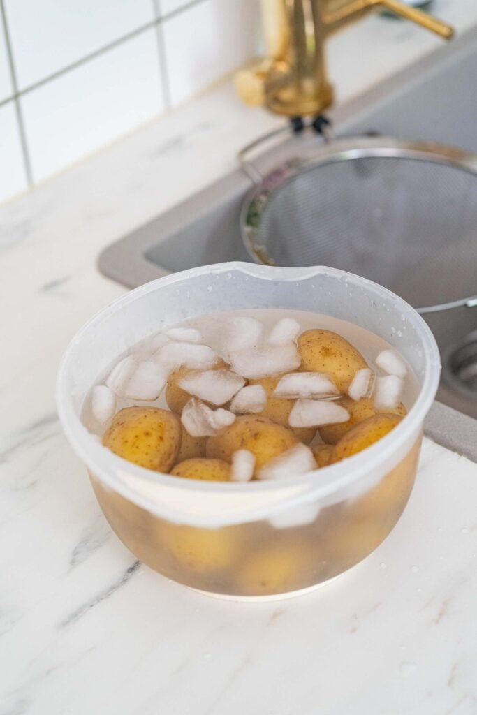 Un bol en plastique rempli d'eau, de glaçons et de pommes de terre jaunes est posé sur un comptoir de cuisine en marbre blanc, près d'un évier équipé d'une passoire, prêt à être transformé en une délicieuse salade de pommes de terre alsacienne inspirée de la cuisine française.