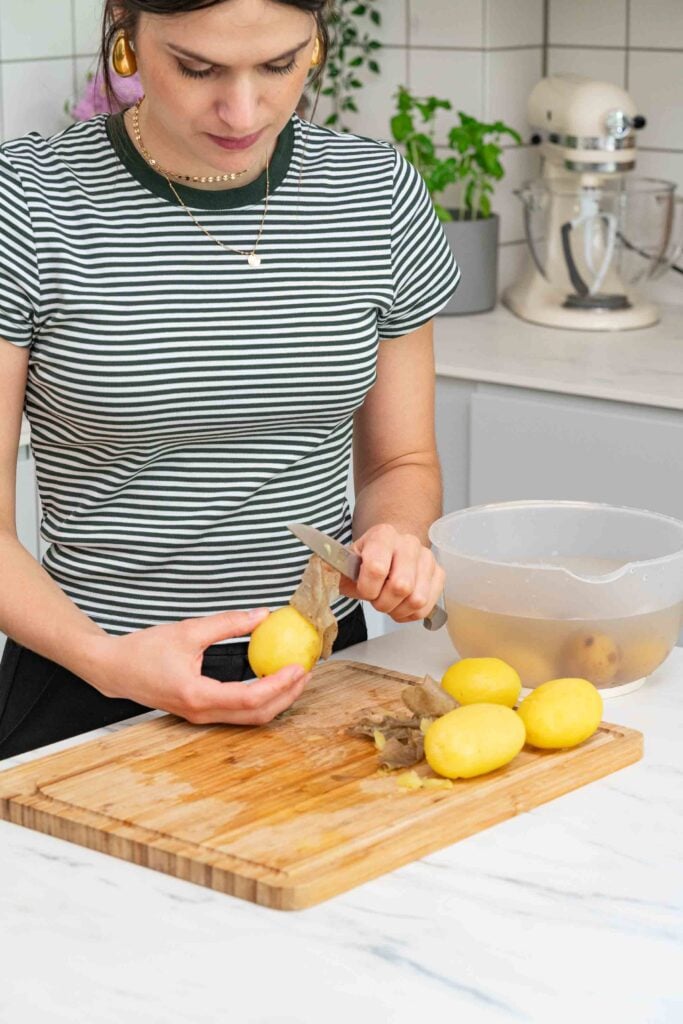 Une personne portant une chemise rayée épluche des pommes de terre sur une planche à découper en bois dans une cuisine, préparant probablement les ingrédients d'une salade de pommes de terre alsacienne. Un bol avec des pommes de terre pelées et des appareils de cuisine sont visibles en arrière-plan, évoquant l'essence de la cuisine française.