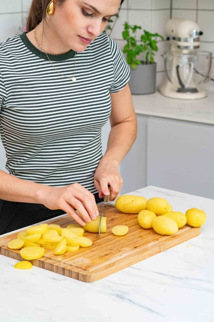 Eine Person in einem gestreiften Shirt schneidet Kartoffeln auf einem hölzernen Schneidebrett in einer Küche, mit einem Mixer und einer Topfpflanze im Hintergrund.