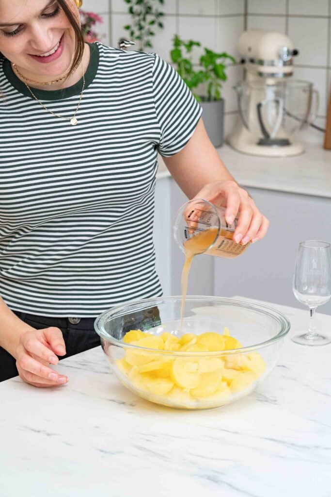 Une personne en chemise rayée verse un liquide d'une tasse à mesurer dans un bol en verre rempli de tranches de pommes de terre, préparant une salade de pommes de terre alsacienne traditionnelle dans la cuisine.