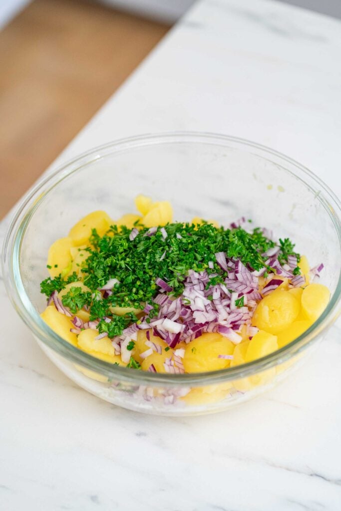 A clear glass bowl filled with chopped yellow potatoes, finely chopped red onions and freshly chopped parsley on a white marble surface.