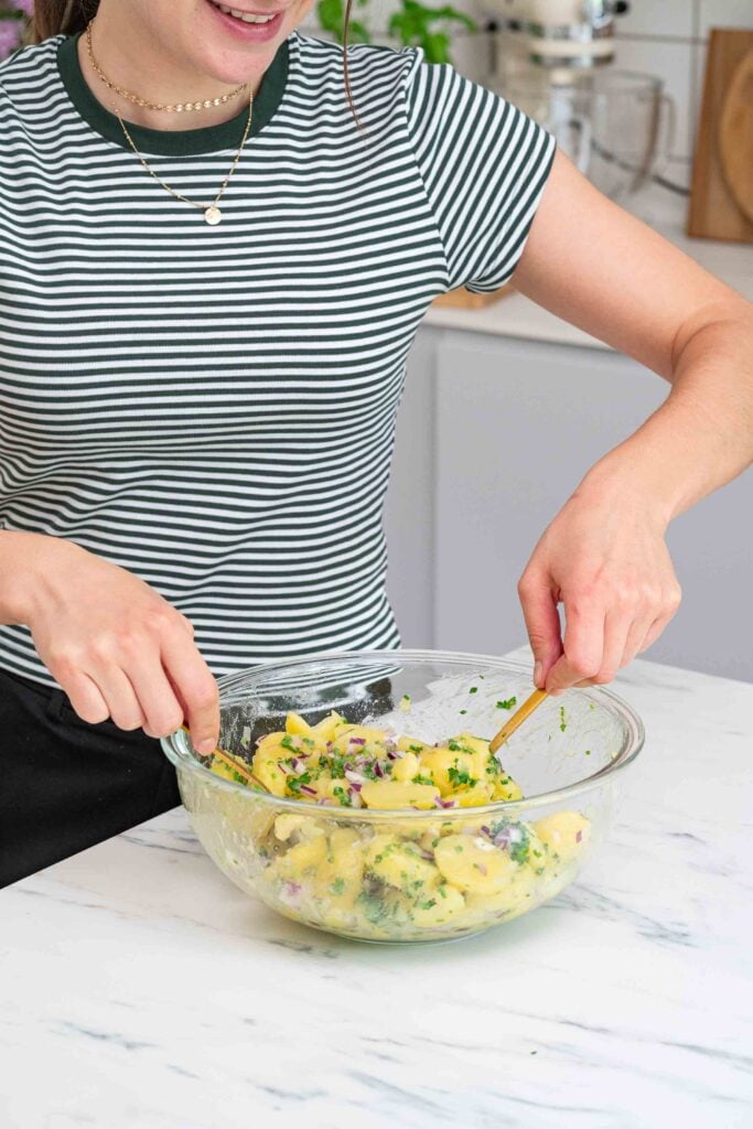 Personne en chemise rayée mélangeant un bol de salade de pommes de terre avec des ustensiles en bois sur un comptoir de cuisine blanc.