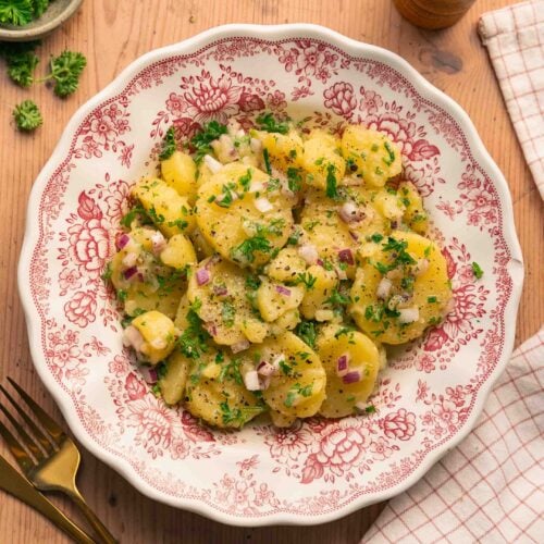 Une assiette avec une salade de pommes de terre alsacienne avec du persil, des oignons rouges et de la vinaigrette, posée sur une table en bois.