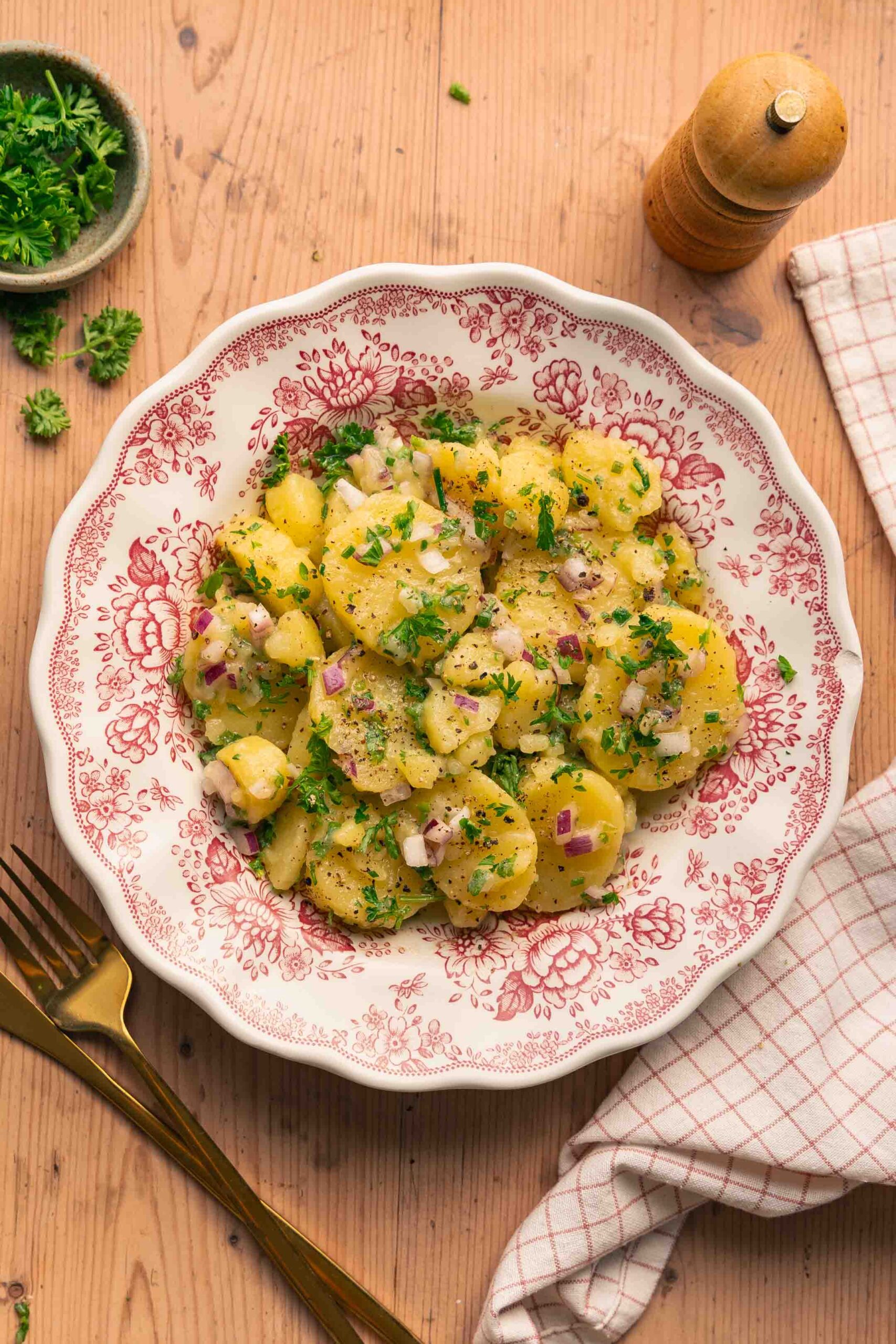 Une assiette avec une salade de pommes de terre alsacienne avec du persil, des oignons rouges et de la vinaigrette, posée sur une table en bois.