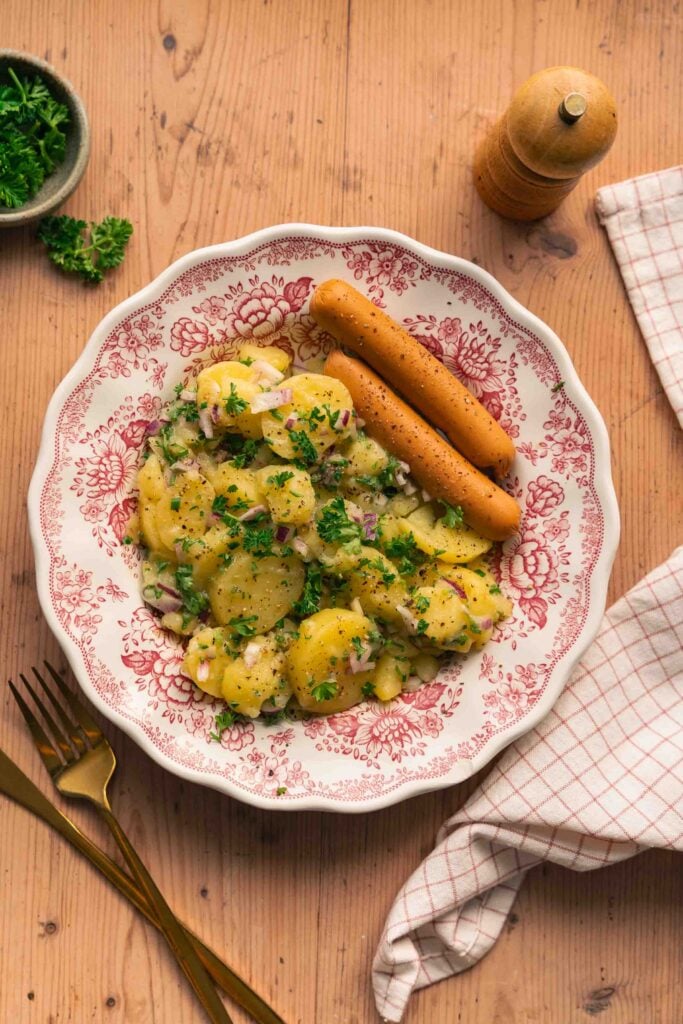 Ein Teller mit elsässischem Kartoffelsalat, garniert mit Kräutern, roten Zwiebeln und begleitet von Grissini. Auf dem Holztisch befinden sich eine Serviette, eine Gabel, ein Messer und eine Pfeffermühle. 