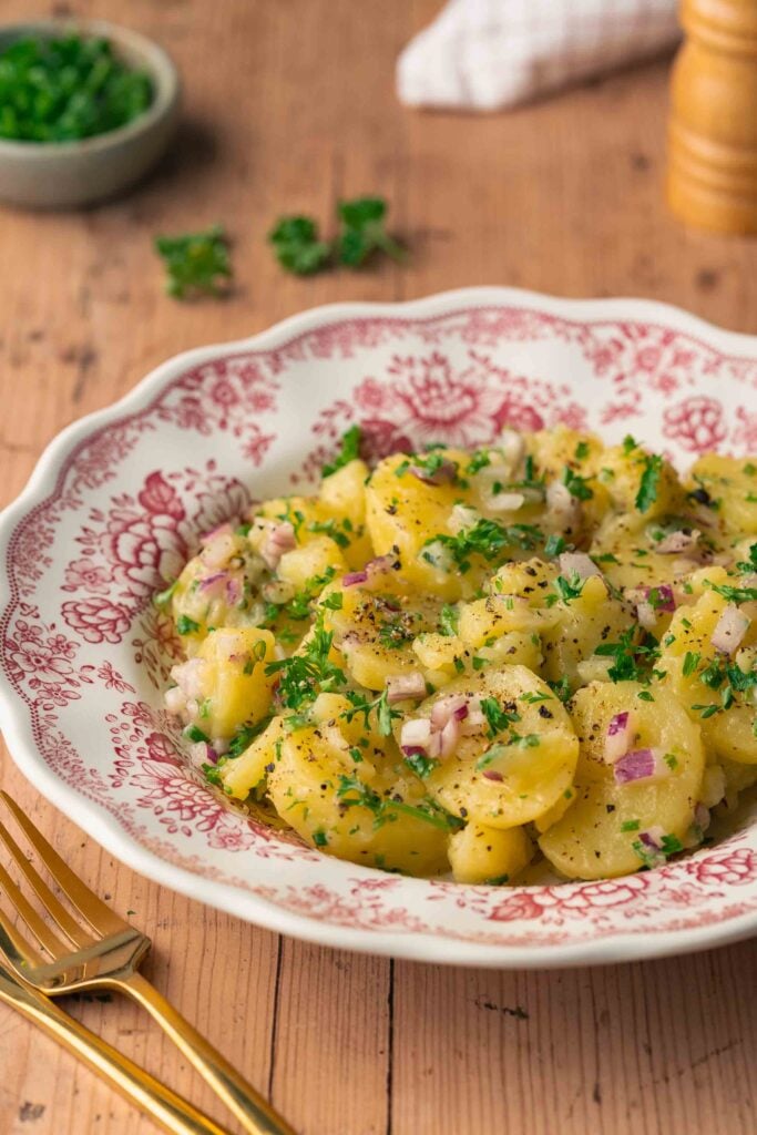 Un bol décoratif rempli de salade de pommes de terre, garni d'oignons rouges hachés et de persil. Une fourchette et un couteau sont placés à côté du bol sur une table en bois.