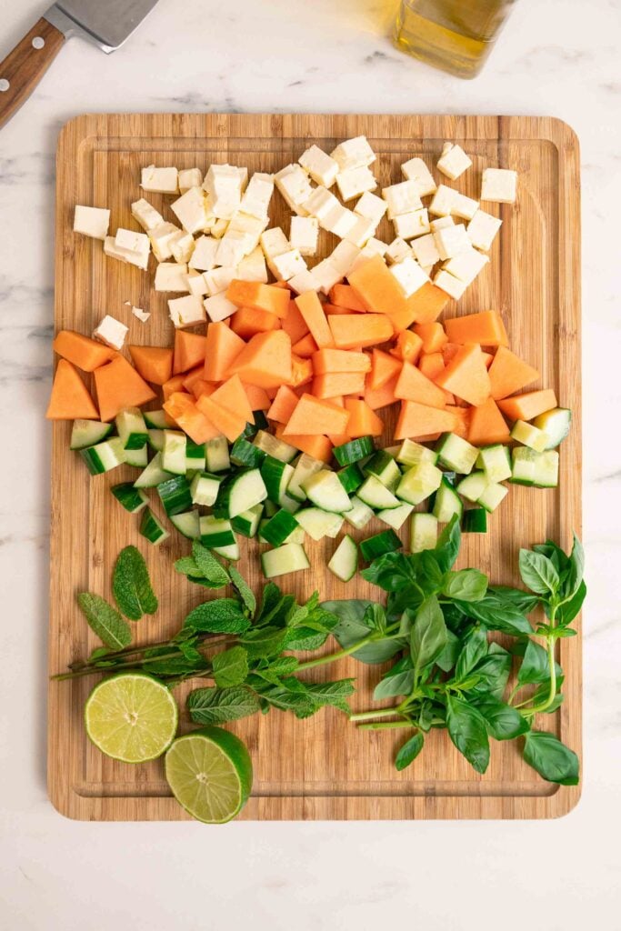 A cutting board with cubes of feta, melon, cucumber, fresh mint leaves, basil leaves and lime halves arranged in rows. A knife and a bottle of olive oil are in the background. 