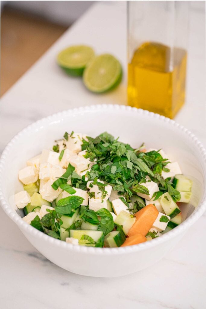 A white bowl filled with a salad of feta cubes, cucumber, melon and chopped herbs. A lime cut in half and a clear bottle of oil are in the background. 