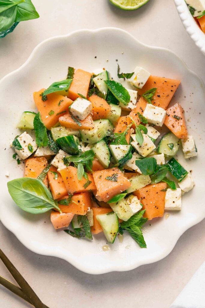 Ein frischer Salat mit Melonenwürfeln, Gurke, Fetakäse und Kräutern, der auf einem weißen Teller serviert wird.